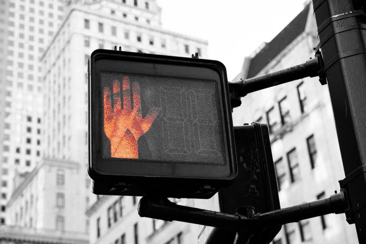 Närbild av trafiksignal som visar orange stopp-handsymbol mot svartvit stadsbild